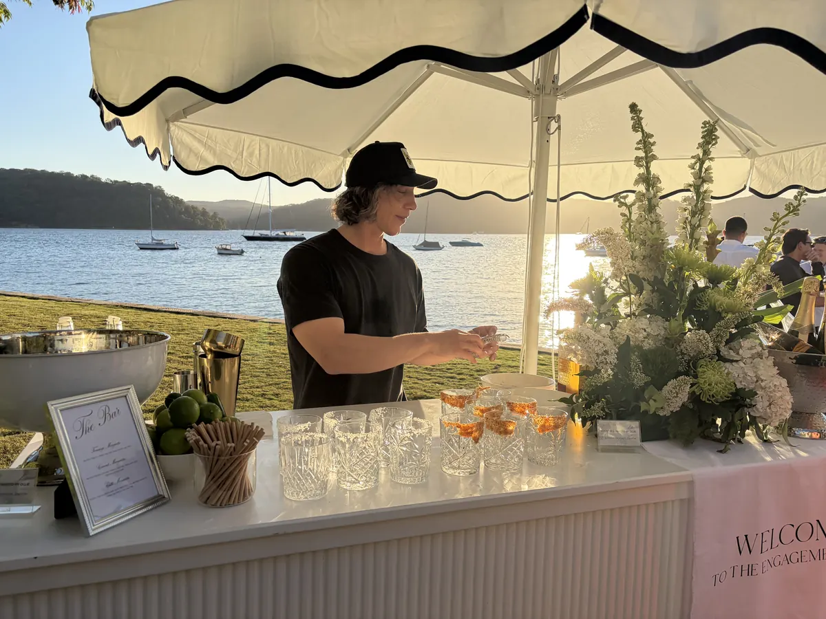 Ollie straining a cocktail behind the mobile bar at golden hour, Pittwater and moored boats behind him