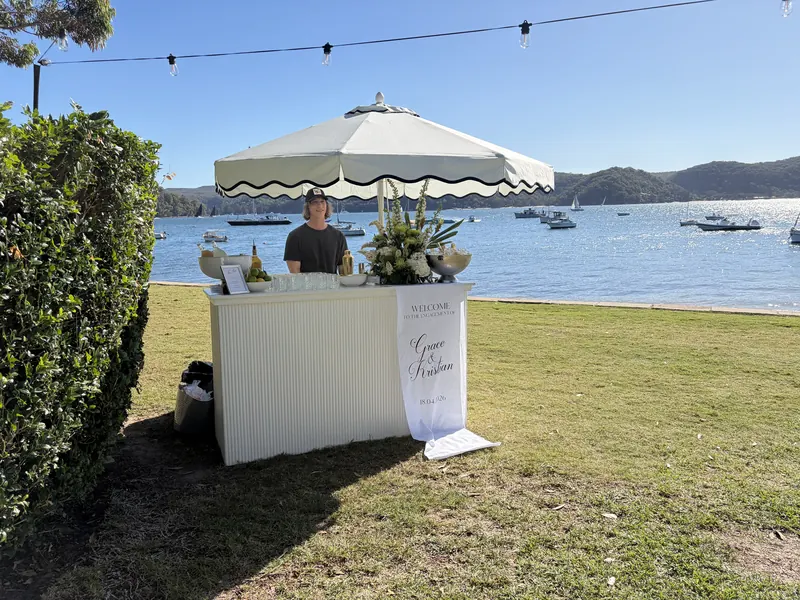 A white fluted mobile bar under a scalloped umbrella on the lawn beside Pittwater